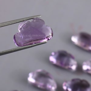 Puede incluir: Primer plano de una piedra preciosa de amatista púrpura tallada en forma de nube. La piedra preciosa está siendo sostenida por unas pinzas. El texto "JEWELSGALLERYBYJYOTI" es visible en la imagen.