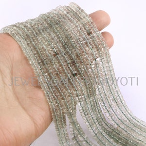 May include: A strand of light green faceted gemstone beads. The beads are small and have a slightly irregular shape. The strand is long and is being held in a hand.