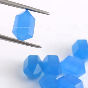 May include: A collection of faceted, light blue gemstones. One gemstone is held by tweezers, while several others are scattered below. The gemstones have a hexagonal shape. The text "JEWELSGALLERYBYJYOTI" is visible on one of the gemstones.