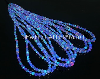 Peut inclure: Plusieurs brins de perles d'opale bleues. Les perles sont sphériques et de tailles variables, créant un effet de cascade. Les perles ont une irisation subtile, avec des éclats de rose et de vert. Le texte "JEWELSGALLERYBYJYOTI" est visible.