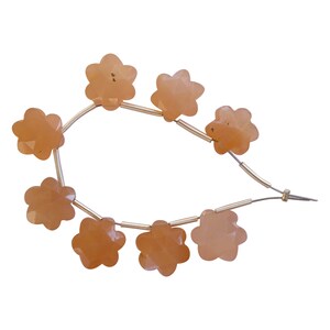 Puede incluir: Hilo de cuentas con forma de flor de color melocotón. Cada cuenta tiene una superficie facetada y está ensartada en un alambre fino de color plateado. Las cuentas están dispuestas en una línea curva sobre un fondo blanco.