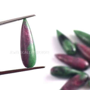Könnte beinhalten: Ein tropfenförmiger Edelstein mit einem grün-rosa Farbmuster. Der Edelstein wird von einer Pinzette vor einem weißen Hintergrund gehalten.