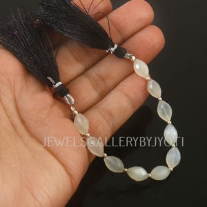 May include: A strand of pale, oval-shaped beads with silver accents and black tassel ends. The beads are strung together, creating a necklace or bracelet component. The text "JEWELSGALLERYBYJYOTI" is visible.
