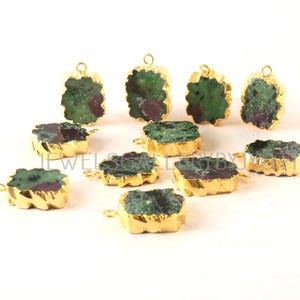 May include: A collection of gold-plated pendants featuring rough-cut ruby zoisite stones. The stones display a mix of green and red hues, with irregular edges. The pendants are arranged on a white surface.