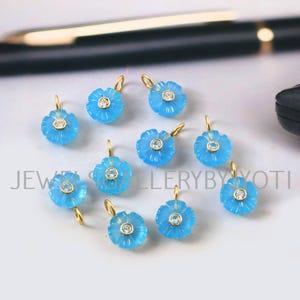 Può includere: Una collezione di pendenti a forma di fiore blu chiaro con anelli dorati. Ogni pendente presenta una piccola pietra rotonda e trasparente al centro. I pendenti sono disposti su una superficie chiara. Il testo "JEWELLERYBYJYOTI" è visibile.