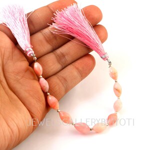 May include: A strand of pink gemstone beads with pink tassels at each end. The beads are oval-shaped and strung together with silver-colored metal. The tassels are made of fine, light pink threads.