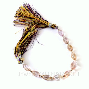 May include: A strand of faceted, oval-shaped beads in shades of pink and peach, strung together. Two decorative tassels with purple, gold, and black threads are attached. The tassels are tied with silver-colored wire. The text "JEWEL GALLERY BY ZOTI" is visible.