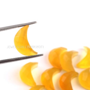 May include: A close-up of a yellow crescent moon shaped gemstone held by tweezers. Several other yellow crescent moon shaped gemstones are scattered on a white surface. The text "JEWELSGALLERYBYJYOTI" is visible in the image.