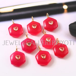 May include: A selection of red, hexagonal pendants with gold loops. Each pendant has a heart-shaped, clear stone in the centre. The pendants are displayed on a white surface, with a black pen in the background.