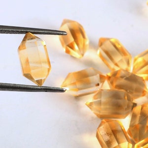 May include: A group of clear, faceted, golden citrine crystals in the shape of double-terminated points. The crystals are arranged on a white surface. One crystal is being held by tweezers.