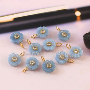 Può includere: Una collezione di ciondoli a forma di fiore blu chiaro, con anelli color oro e una pietra trasparente al centro. I ciondoli sono sparsi su una superficie rosa chiaro, con una penna e un oggetto nero sullo sfondo. Ideali per la creazione di gioielli.
