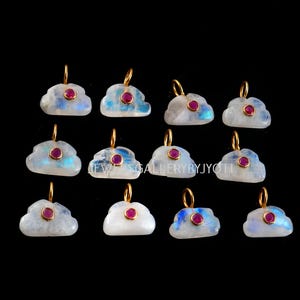 May include: A collection of cloud-shaped pendants with gold-colored loops. Each pendant features a white or blue-tinged stone with a small, round, red gemstone in the center. The pendants are displayed against a black background.