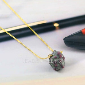May include: A gold-colored chain necklace with a rough, irregularly shaped pendant. The pendant is a mix of green and red hues. The necklace is displayed on a light-colored surface. The text "JEWELSGALLERYBYJOH" is visible.