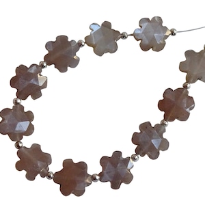 Puede incluir: Una hebra de cuentas de piedras preciosas de color marrón claro, cada cuenta tiene forma de flor con múltiples puntas. Las cuentas están enhebradas en un cordón plateado con cuentas espaciadoras plateadas.