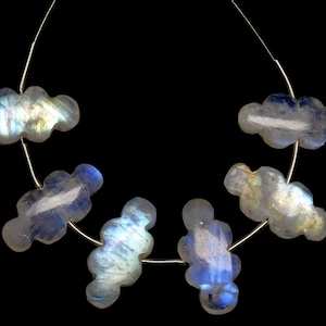 May include: Six cloud-shaped moonstone beads strung on a silver wire. The beads have a translucent, iridescent quality with blue and white hues. The beads are arranged against a black background.