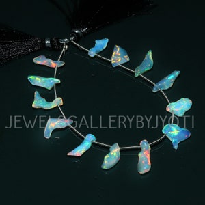 May include: A strand of opal gemstone beads. The beads are in irregular shapes, displaying a mix of blue, white, and iridescent colours. They are strung on a thin silver wire. The background is a dark, reflective surface. The text "JEWELS GALLERYBYJYOTI" is visible.