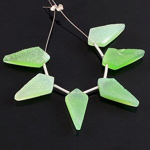 Puede incluir: Una hebra de cuentas de piedras preciosas verdes con un alambre de plata enhebrado a través de ellas. Las cuentas son facetadas y tienen una superficie rugosa y texturizada.