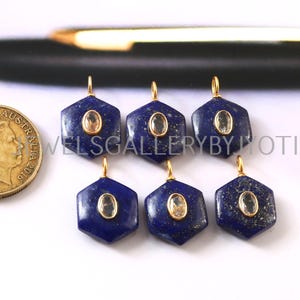 Op de afbeelding: Zes zeshoekige lapis lazuli hangers met goudkleurige beugels en een centrale ovaalvormige steen. De hangers zijn diepblauw met gouden vlekken. Een gouden munt is zichtbaar. De tekst "JEWELS GALLERY BY JOTI" is zichtbaar.