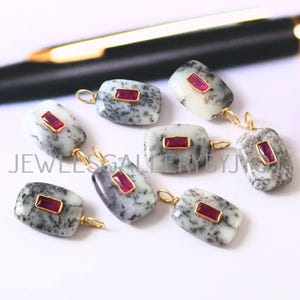 May include: Seven rectangular pendants with a gold loop and a small, rectangular, red gemstone set in a gold frame. The pendants have a white and gray speckled pattern. The text "JEWELS GALLERY BYJ" is visible.