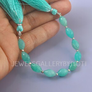 May include: A strand of turquoise-colored gemstone beads with a tassel. The beads are oval-shaped and strung together with small silver-colored beads. The tassel is made of turquoise-colored thread. The text "JEWELSGALLERYBYJYOTI" is visible.