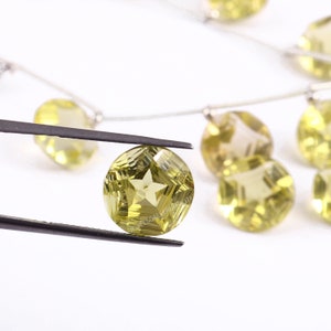 May include: Close-up of a faceted, light yellow gemstone held by tweezers. The gemstone has a star-shaped design in the center and is part of a strand of similar stones. The background is white, highlighting the gems.