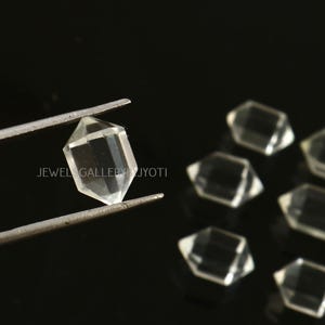 May include: A group of clear quartz double terminated points, also known as double pointed crystals, are displayed on a black surface. One point is held in tweezers.  JEWEL GALLERY BY JYOTI is printed on the surface.