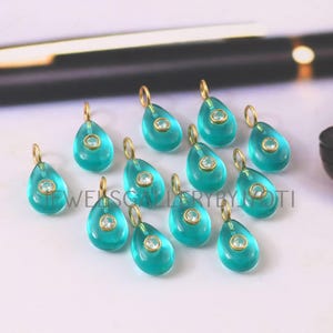Puede incluir: Un conjunto de colgantes turquesas en forma de lágrima con aros dorados. Cada colgante tiene una pequeña piedra redonda y transparente en el centro. Los colgantes están dispuestos sobre una superficie blanca. Texto en la imagen: JEWELSCALLERYBYJYOTI.