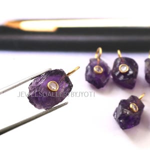 May include: Raw amethyst pendants with gold-colored bails and small, round, clear stones set in the center. The pendants are held by tweezers. The text "JEWELLSGALLERYBYJYOTI" is visible.