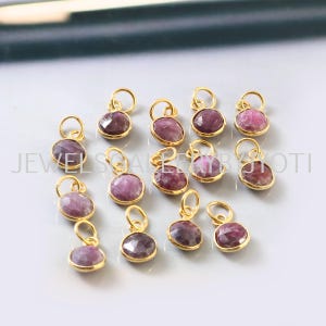 May include: A collection of 14 gold-plated charms featuring round, faceted, pink gemstones. The charms have a loop at the top for attaching to jewellery.
