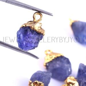 May include: A close-up of a purple gemstone pendant with a gold-plated setting. The pendant is in the shape of a rough, natural crystal. The pendant is being held by a pair of tweezers.