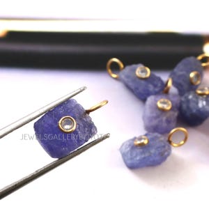 May include: Close-up of several rough-cut, blue-violet tanzanite gemstones with gold-colored loops. One stone is held by tweezers. Each stone has a small, round, clear gem set in a gold bezel. The text "JEWELSGALLERYBYIYOT" is visible.