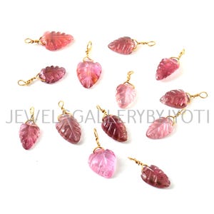 Könnte beinhalten: Eine Sammlung von rosafarbenen, blattartigen Edelsteinanhängern mit goldfarbenen Drahtschlaufen. Die Anhänger sind auf einer weißen Oberfläche verstreut.