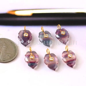May include: Six leaf-shaped pendants with gold-colored loops and a small, round, clear stone in the center. The pendants are purple, pink, and clear. A coin is in the background.