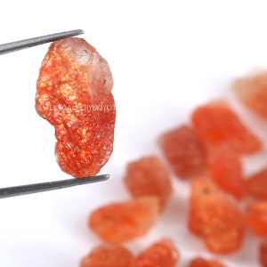 Op de afbeelding: Een close-up van een ruwe, oranje sunstone edelsteen met een metallic glans. De steen wordt vastgehouden door een pincet en is omringd door andere kleinere sunstone edelstenen.