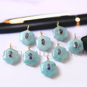 Può includere: Una collezione di pendenti con pietre preziose acquamarina. Ogni pendente esagonale presenta un'ansa dorata e una pietra ovale blu scuro al centro. I pendenti sono di colore azzurro chiaro e sono esposti su una superficie bianca.