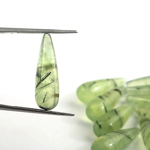 以下が含まれることがあります： 黒いインクルージョンが入った緑色の宝石の涙型ビーズの集まり。ビーズはピンセットで保持され、積み重ねられています。