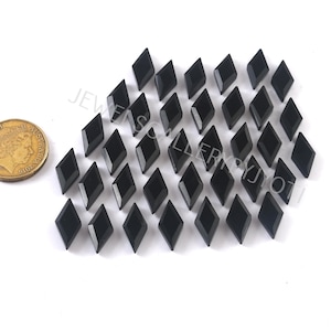 May include: A collection of black, diamond-shaped gemstones. The gemstones are faceted and have a glossy finish. A gold coin is in the image for size comparison. The gemstones are arranged on a white surface.