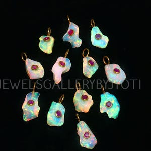 Può includere: Una collezione di ciondoli in opale con anelli color oro e piccole gemme rosse rotonde. Gli opali hanno un aspetto iridescente colorato, con sfumature di blu, verde e rosa. Il testo "JEWELSGALLERYBYJYOTI" è visibile.