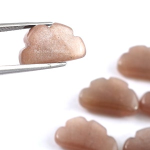 Pode incluir: Cinco cabochões de pedra do sol esculpidos em forma de nuvens. As pedras são de cor pêssego claro com um acabamento brilhante e iridescente.