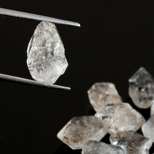 May include: A clear, faceted, rough-cut diamond held by tweezers. Several other similar diamonds are scattered on a black surface.