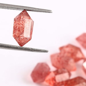 May include: A collection of red quartz crystals, some of which are held by tweezers. The crystals are faceted and have a translucent appearance.