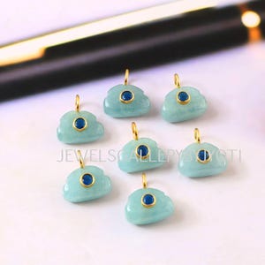 Puede incluir: Siete colgantes en forma de nube azul claro, cada uno con una pequeña piedra central azul oscuro y un lazo dorado. Los colgantes están dispuestos sobre una superficie blanca. El texto "JEWELSCALLERYJYOTI" es visible.
