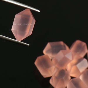 May include: A close-up of a pink rose quartz crystal, with a faceted, octagonal shape. The crystal is held by tweezers, and several other rose quartz crystals are visible in the background.