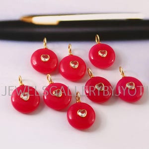 May include: Eight red, circular pendants with gold loops. Each pendant has a heart-shaped, clear gemstone in the centre. The pendants are presented on a light-coloured surface.