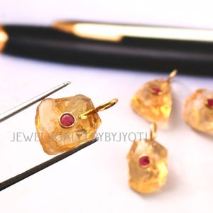 Puede incluir: Primer plano de colgantes de piedras preciosas en bruto, de color amarillo dorado, con una pequeña gema roja redonda en el centro. Cada colgante tiene un lazo dorado para una cadena. El texto "JEWEL GALLERYBYJYOTI" es visible.