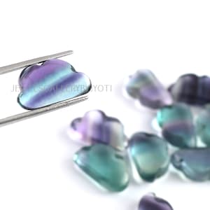 May include: A group of small, iridescent, cloud-shaped fluorite stones in shades of purple, blue, and green. The stones are arranged on a white surface.