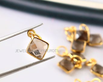 May include: A gold-colored pendant with a square, smoky-colored gemstone is held by tweezers. The pendant has a small loop at the top. Other similar pendants are visible in the background.
