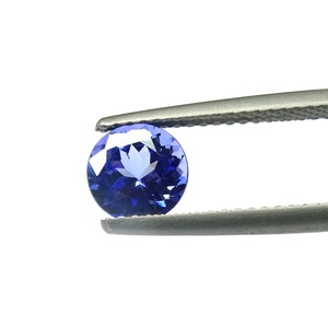 Peut inclure: Une pierre précieuse ronde de couleur bleue, tenue par des pinces. La pierre précieuse a un centre bleu clair et un bord extérieur bleu foncé.