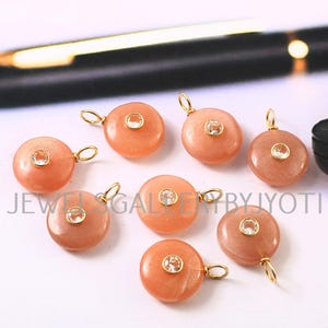 May include: A collection of eight peach-colored gemstone pendants with gold-colored loops. Each pendant features a small, clear, round stone in the center. The pendants are arranged on a white surface, with a pen in the background.