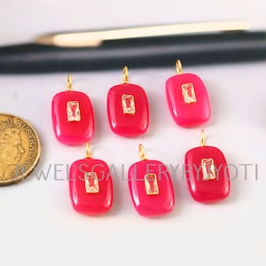 Peut inclure: Six pendentifs rectangulaires rose vif avec des boucles dorées et une pierre rectangulaire transparente au centre. Les pendentifs sont disposés sur une surface blanche. Une pièce d'or et un stylo sont également visibles.